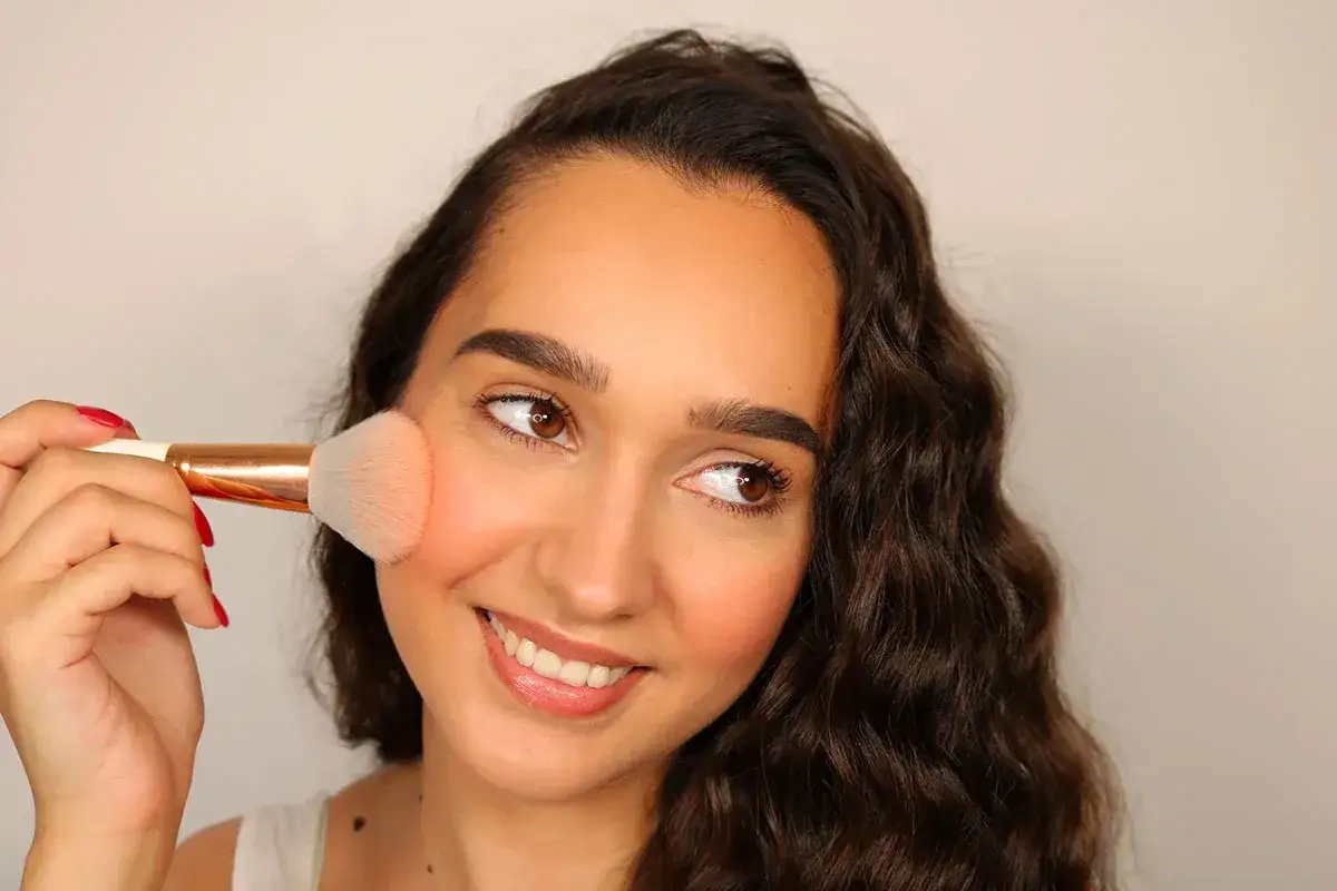Donna sorridente applica blush o fard sulle guance con un pennello, creando un effetto naturale e radioso.