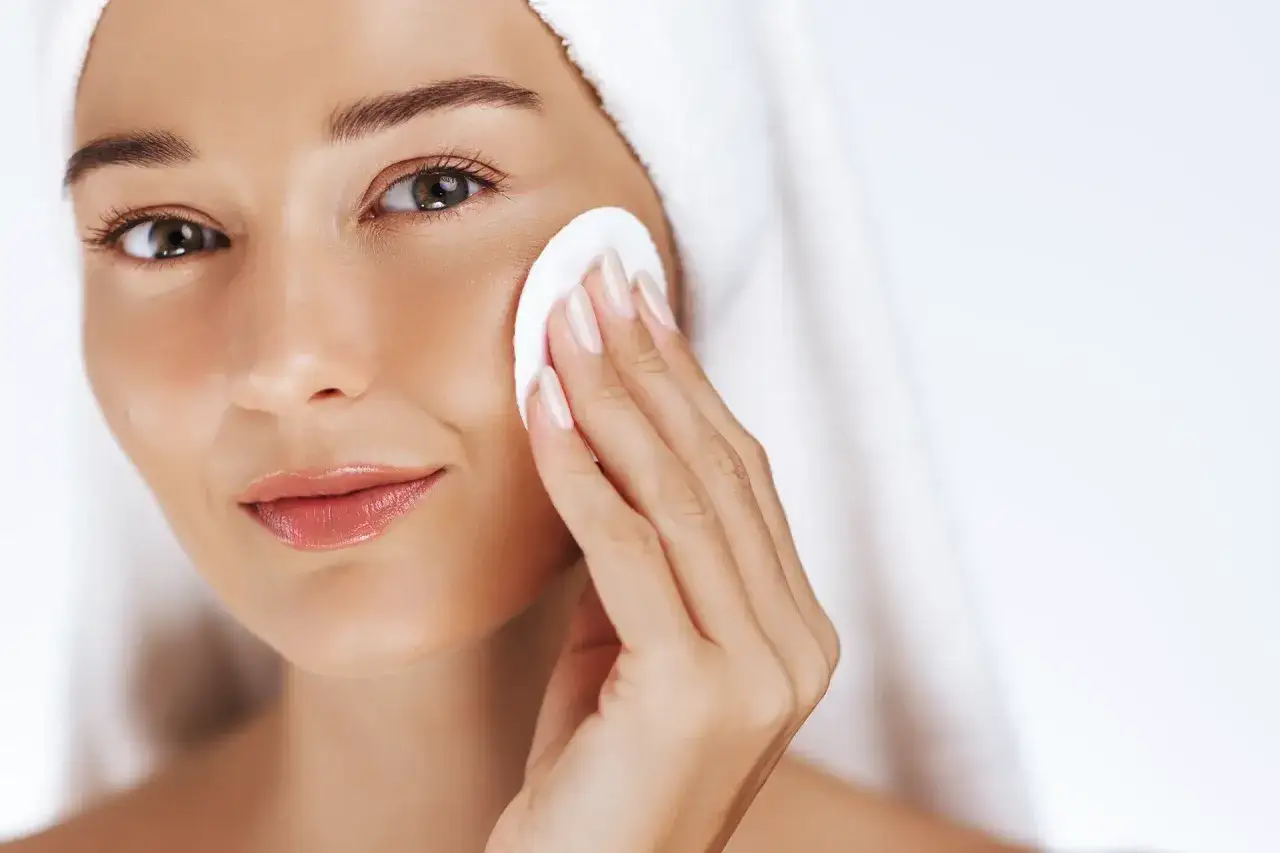 Donna con asciugamano in testa si pulisce il viso con un dischetto di cotone, un gesto essenziale per capire come fare la skincare.