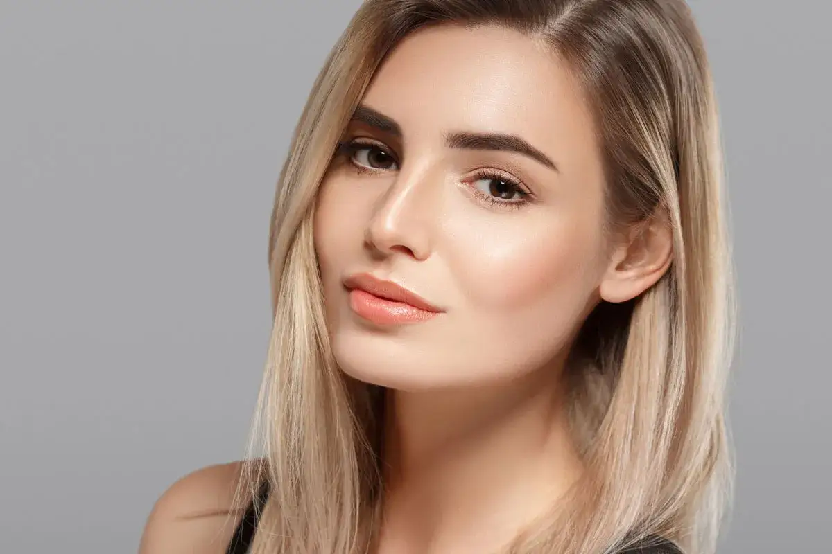 Close-up of a woman's face with natural makeup and blonde hair against a gray background.