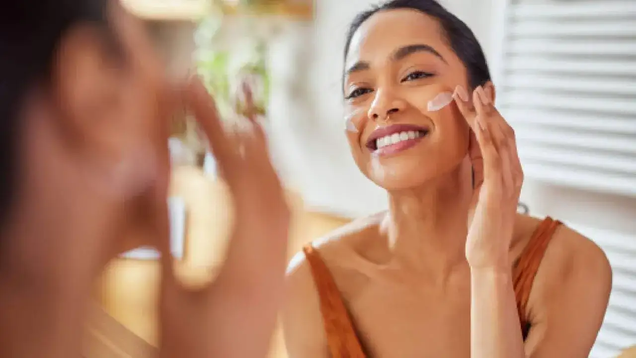 Donna sorridente applica crema viso con tocchi leggeri sulle guance, un gesto quotidiano per una pelle radiosa.