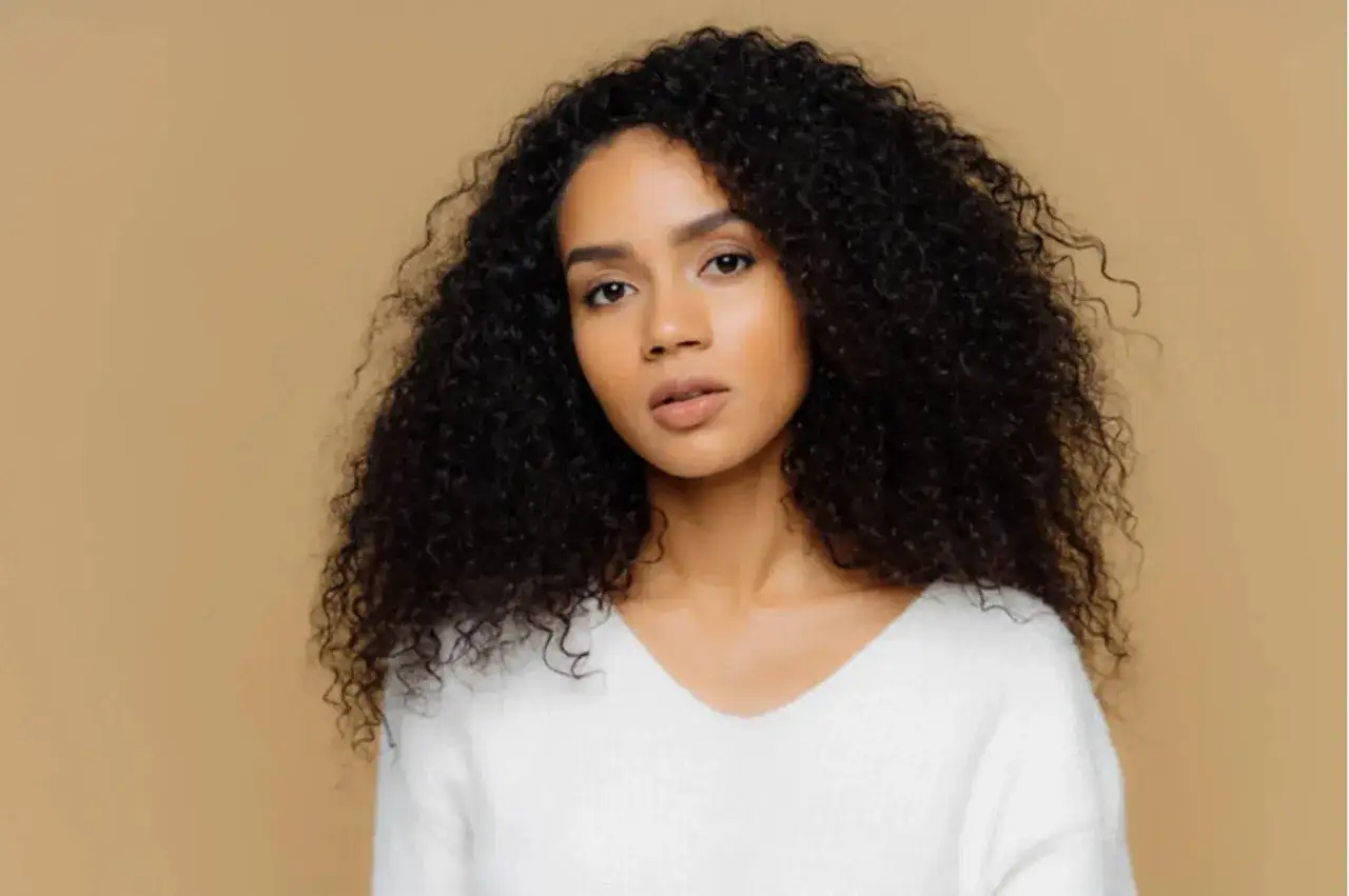 Donna con splendidi capelli ricci secchi, voluminosi e scuri, indossa un maglione bianco.